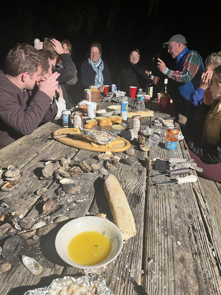 The family around a long weathered table covered in oyster shells at a nighttime roast